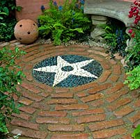 Circular courtyard with stone bench and ferns surrounding tiled floor with central star motif created from white and grey pebbles.