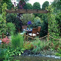 Wooden pergola clothed in fragrant Trachelospermum jasminoides, beside pool edged in sage, hosta, astilbe and grasses. Dining area enclosed in evergreen shrubs.