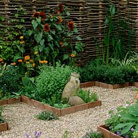 Woven hazel screens create privacy around a small courtyard garden with gravel paths and raised beds of herbs, box and perennials edged in terracotta tiles laid on edge.