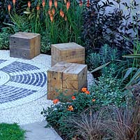 Tranquil meditation garden with individual oak cube seats set around circular pebble mosaic. Orange and maroon plants: kniphofia, rose, elder and sedge. Thyme path.