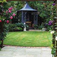 Pergola clad in Rosa Handel frames view of small town garden with brick-edged, circular lawn and grey-painted summerhouse between hostas and trees.