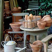 Collection of terracotta flower pots in a wire basket on table, watering can and old chair in working area.