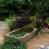 Water lilies grown in shallow, stone trough set on gravel amidst grasses, cotinus, fern and astrantia.
