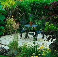 In small suburban garden, a secret courtyard with table and chairs, hidden behind cercis, tulip tree, phormium, grasses and laurel hedge.
