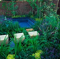 Wood oak cubes form stepping stones through planting and across small wildlife pool beside sunken, slate patio with wall seats for relaxing amongst aromatic plants.