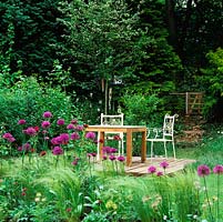Small wooden deck with table and chairs set beside path leading to gate opening into woodland, glimpsed through Allium 'Purple Sensation'