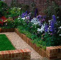 Gravel path edged in low brick wall containing raised bed of delphinium, mallow and pinks. Behind: woven willow fence panel hurdle.