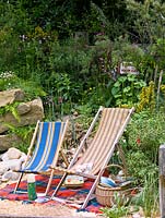 Seaside-themed garden with boulders retaining a bank of wildflowers, deckchairs and picnic basket on rug on sand.