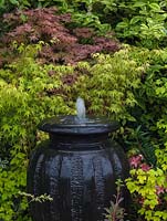 Huge ceramic urn has bubble fountain cascading water over its sides, against backdrop of maples.