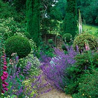 Gravel path lined in Nepeta 'Six Hills Giant', eremurus, foxglove, cistus, box, holly topiary and Italian cypress leads to old wooden gate in dry stone wall.
