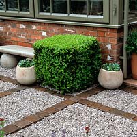 Path of stone chippings on membrane, contained by bricks, forming large rectangles. Box topiary in cube beside stone seat and brick wall of conservatory.