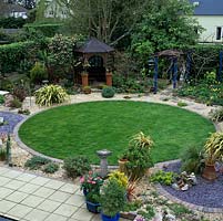 Town garden based on 3 linked circles - 2 slate beds by a brick-edged, circular lawn, edged by winding, gravel path. Gazebo. Pergola. Evergreen phormium, camellia, grasses.