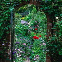 View through gate to Lutyens-style bench in herb garden. Beds of poppy, aquilegia, clematis, rose, hardy geranium and herbs punctuated by paths and box balls.