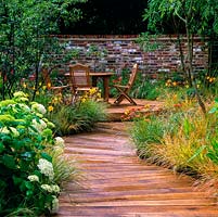 Seating area with table and chairs, on raised deck beneath canopy of birches. Curving timber path edged in grasses, hydrangea, daylily and achillea.