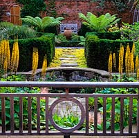 Oak balustrade overlooks sunken pool with astilbe, primula and hosta. Edged by Ligularia przewalskii. Beyond, yew hedges and sunlit tree ferns. Pot in rose bed and seat.
