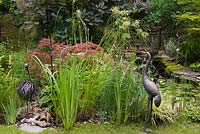 Metal ostrich and heron bird sculptures with Acer palmatum inabe-shidare - Japanese Red Maple tree next to pond with Iris 'Yellow Flag' in backyard garden in summer, Quebec, Canada
