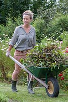 Woman pushing wheelbarrow with pruned shrubs and flowers to the compost heap