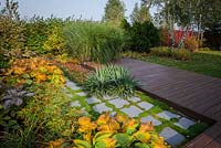 Square brick path surrounded by blue hosta sieboldiana. View of wooden terrace and long border created by grass, conifers, and red cornus twigs
