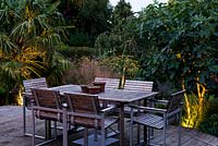 A raised wooden deck with garden furniture for outdoor dining. Uplighters illuminate surrounding planting including Trachycarpus fortunei palm, Deschampsia cespitosa 'Bronzeschlier' 