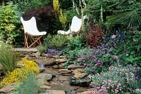 Patio with two white canvas chairs. Artificial stream. Acer, sambucus, cytisus, genista, osteospermum, verbascum. 