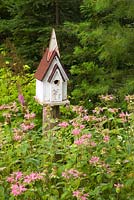Wood and metal birdhouse in border planted with pink Monarda flowers against a backdrop of Picea - Spruce and Pinus - Pine tree foliage in backyard country garden in summer, Quebec, Canada