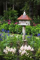 Border with wood and metal birdhouse and planted with pink and white Astilbe, blue Delphinium flowers in front yard country garden in summer, Quebec, Canada