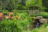 Border planted with orange Hemerocallis - Daylily, burgundy red Astilbe X arendsii 'Burgunderrot' next to natural stone man-made waterfall in front yard country garden in summer, Quebec, Canada