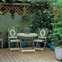 Chair and tables on shady wooden deck, hidden behind fence panels, pots and Trachelospermum jasmoides, tucked away at bottom of 27m x 13m town garden.