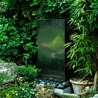 Stainless steel block water feature, set on bed of pebbles, dimly reflects rockery beyond.