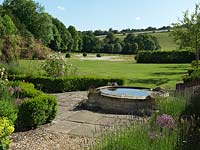 An old stone pool surrounded by formal buxus hedges, Lavandula Hidcote and Allium Christophii. Beyond is a large lawn with a wildflower area and views of the hampshire hills.