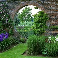 Circular moongate in old brick wall, flanked by roses, magnolia, iris and aquilegia, frames view of parkland, cows and distant countryside beyond.