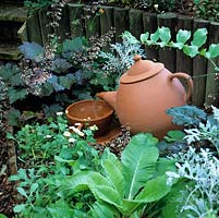 Pouring terracotta tea pot water feature placed on pebbles in shady corner.