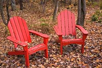 Two old red painted wooden adirondack chairs in a deciduous tree forest of Acer. Laurentians, Quebec, Canada