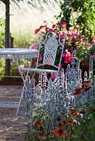 An ornate French circular white table and  matching chair at the end of a gravel path.  With Helenium Moerheim Beauty and Stachys Byzantina. At Domaine de Cambou, Verfeil, Haute-Garonne, Midi Pyranees, France.