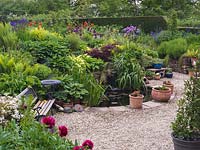 Seen over red peony, bank of oriental poppy, Iris sibirica, centaurea, fern, allium, hosta, acer and  bergenia edge pool, bench and sunken terrace. 