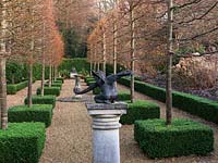 In secret, sunken garden, avenue of pleached limes, stripped of leaves and with their trunks encased in box squares. On pedestal, statue of Cupid. Central sundial.