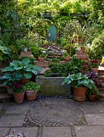View of 30m x 8m back garden. Patio - old water tank and pots of hostas, holly and pelargonium - leads to wisteria clad pergola, past raised beds of allium, a greenhouse, to door in back wall.