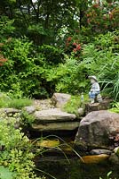 Sculpture of young boy fishing on the edge of a pond with Alchemilla mollis - Lady's mantle, Typha latifolia - Common Cattails and red flowering Weigela shrubs in backyard country garden in summer, Quebec, Canada