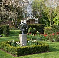 A formal grass parterre surrounded by yew - Taxus hedging. An Armillary sphere is set in boxed edged bed with Narcissus and the bed behind is planted with Tulips and roses.