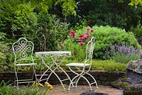 White cast-iron metal table and chairs on paving stone patio with red Lilium Asiatic 'America' - Lily flowers and Pinus strobus 'Blue Shag' - Pine shrub, Lavandula angustifolia 'True Blue' - Lavender in raised stone border in backyard country garden in summer, Quebec, Canada