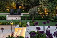 Town garden designed by Kate Gould, lit at night. Flames from an open gas fireplace illuminate the sunken terrace which is edged in beds of box balls interspersed with purple and white allium.