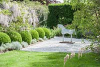 Galvanised metal horse beside pergola draped with flowering wisteria and framed by clipped box balls. 