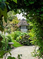 A thatched summer house and bog garden seen from under a dense foliage arch.