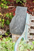 Decorative carved slate with birds beside bench in front garden. 