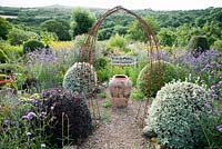 A large oil jar marks the centre of the Rickyard, framed by rusty metal 'arches' and pots of Aeonium 'Zwartkop' with scarlet pelargoniums. Gravel path is lined with lavender, Verbena bonariensis and clipped pittosporums, hebes and box. Dyffryn Fernant, Fishguard, Pembrokeshire, Wales, UK