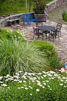 Top view of flagstone patio with blue bench, brown metal table and chairs next to man-made pond and borders planted with white Leucanthemum vulgare - Oxeye Daisy flowers and Miscanthus sinensis - Ornamental Grass in private front yard country estate garden in summer, Quebec, Canada