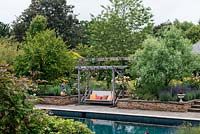 A view across a swimming pool to a swing seat between Cornus kousa 'Chinensis and Fraxinus angustifolia 'Raywood'.