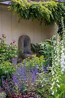 A stone wall mounted water feature behind a border planted with Digialis 'Alba', Alchemilla mollis, heuchera and Nepeta 'Six Hills Giant'.