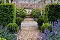 A formal walled garden with gravel path leading to a water feature. The path is lined by box balls in containers, yew hedging, cat mint and choisya.
