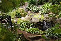 Man-made creek and cascading waterfall bordered by Hosta and Lysimachia nummularia 'Aurea' - Golden Creeping Jenny plants in backyard garden in summer, Quebec, Canada